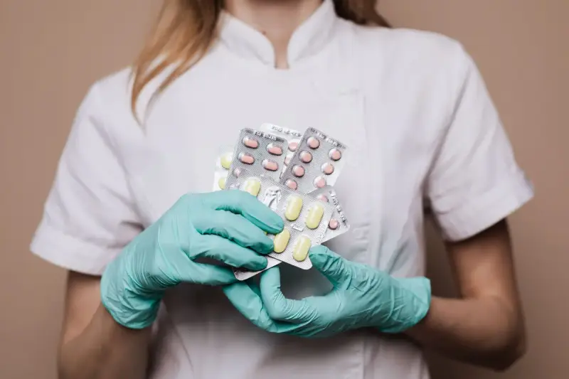 Gloved hands of a pharmacy worker in a white tunic holding several blister packs of tablets
