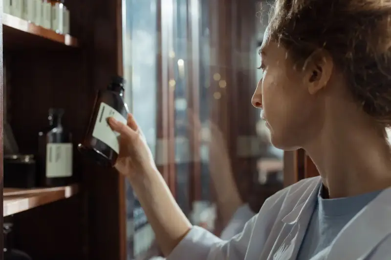 Pharmacist in a white coat taking a labeled brown bottle from a wooden shelf