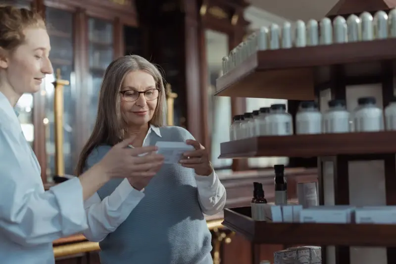 Pharmacist in a white coat helping a smiling older woman read a product label beside the shelves