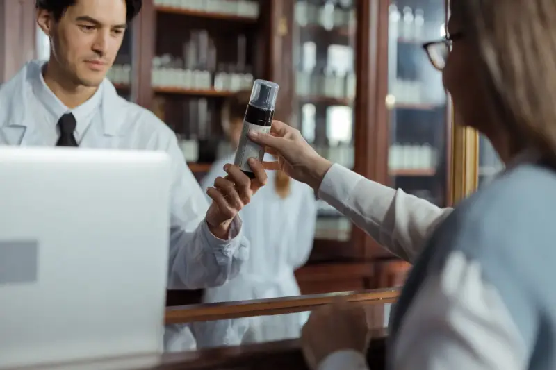 Pharmacist at a laptop handing a bottle across the counter to a customer
