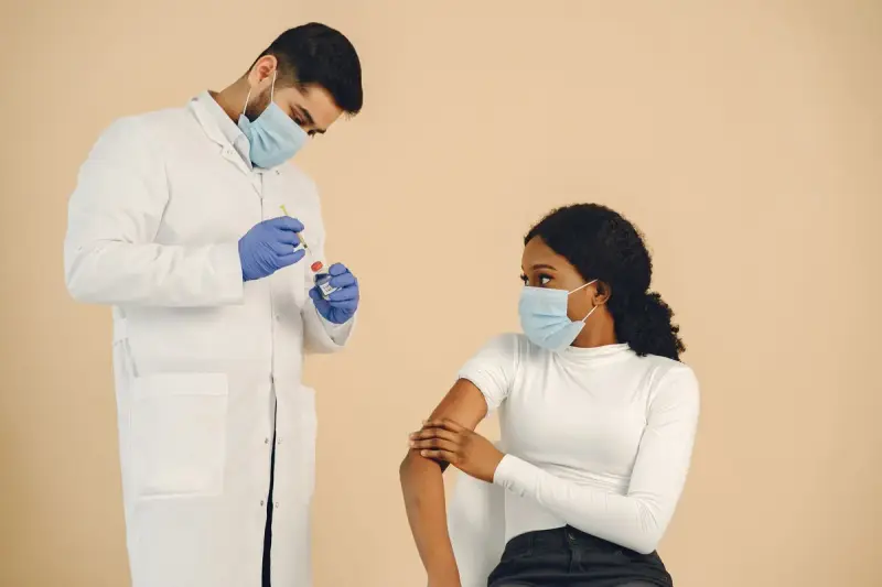 Masked clinician in a white coat preparing a vaccine syringe beside a seated young woman