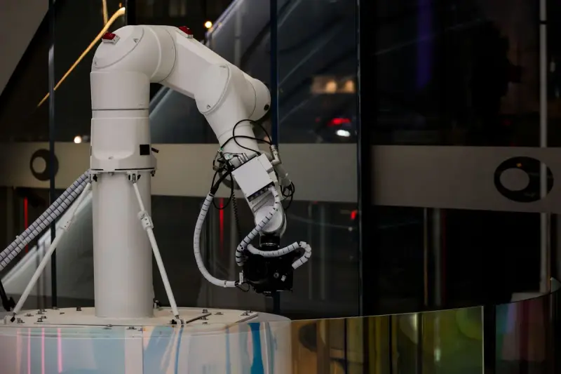 White six-axis robotic arm with cabled gripper mounted on a pedestal against glass walls