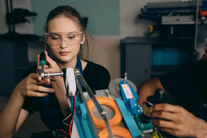 Young woman in safety glasses assembling a small robotic device at a workbench