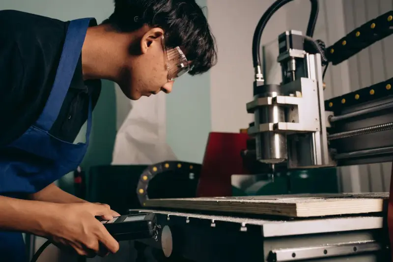 Machinist in safety glasses running a benchtop CNC mill with a handheld controller