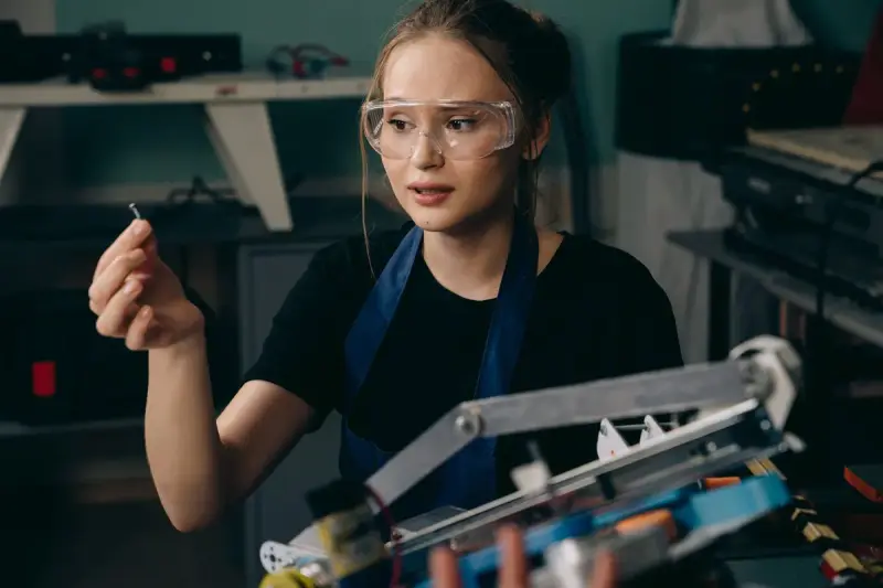 Engineer in safety goggles holding up a small metal pin beside a robotic arm assembly