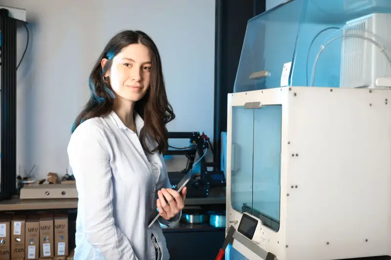 Engineer holding a tablet beside an enclosed 3D printer in a prototyping workshop