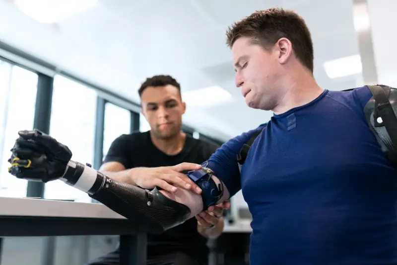 Engineer adjusting a carbon-fiber bionic arm on a man's forearm at a lab bench