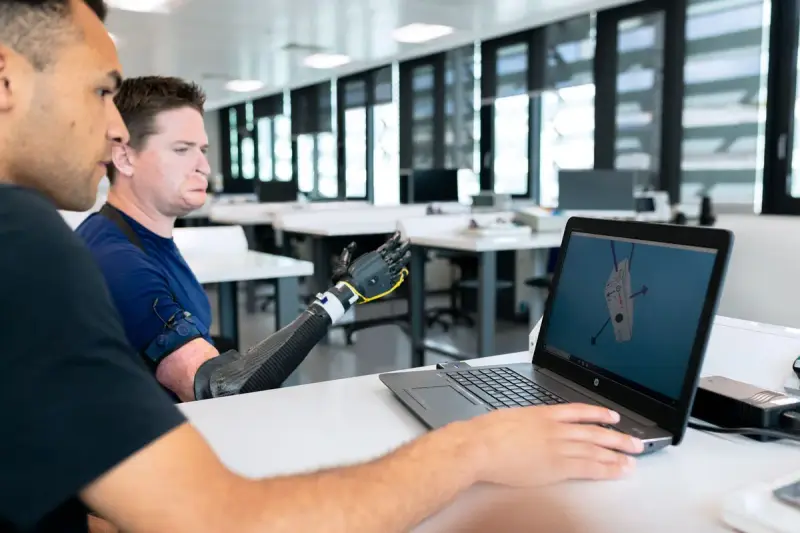 Engineer at a laptop reviews a design model while a colleague tests a prosthetic arm in a bright lab