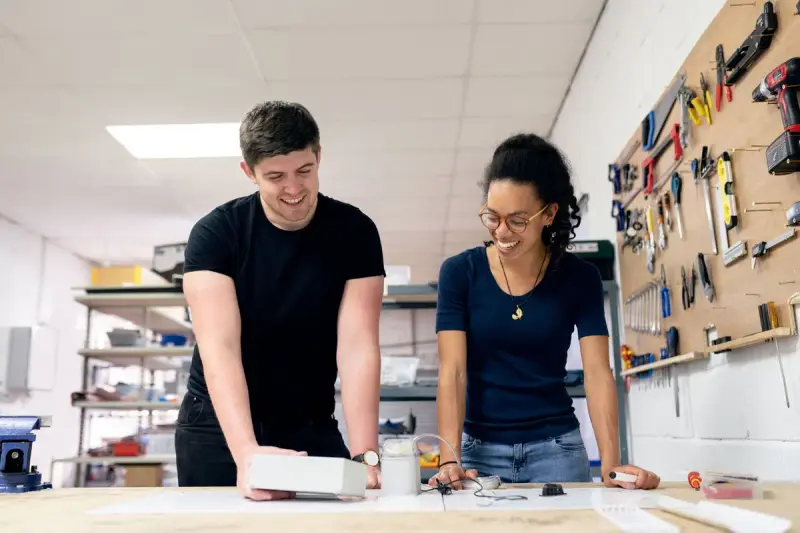 Two smiling engineers, a man and a woman, test a small prototype device on a workshop bench
