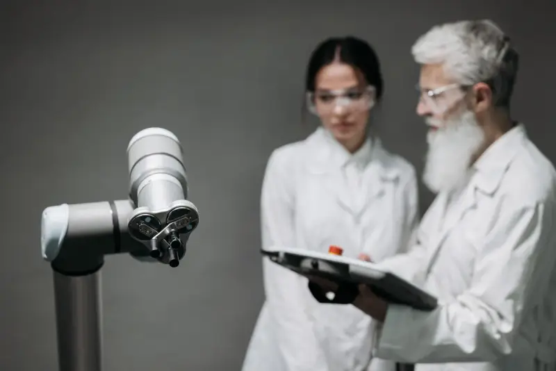 Two researchers in lab coats and safety glasses review a tablet beside a robotic arm