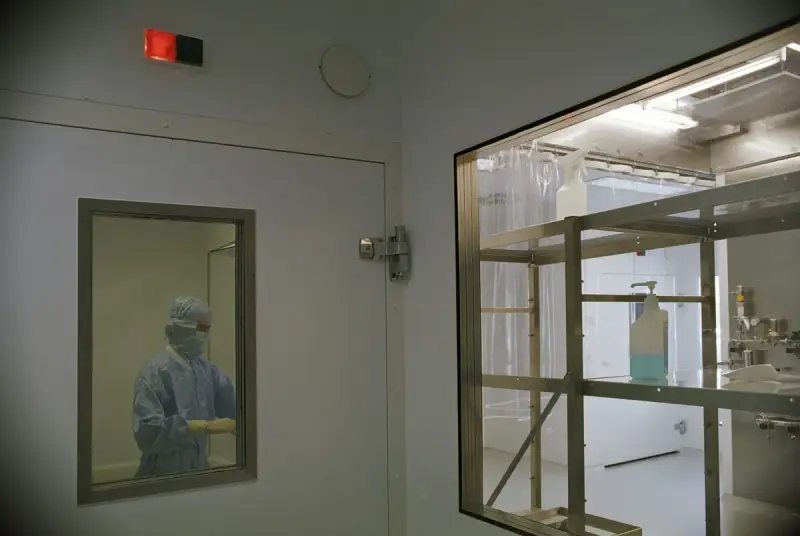 Technician in a full cleanroom suit seen through a door window beside a pass-through cabinet