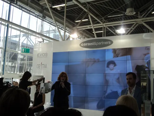 A woman presenting beside a live demonstration and large video wall at Cosmoprof Bologna