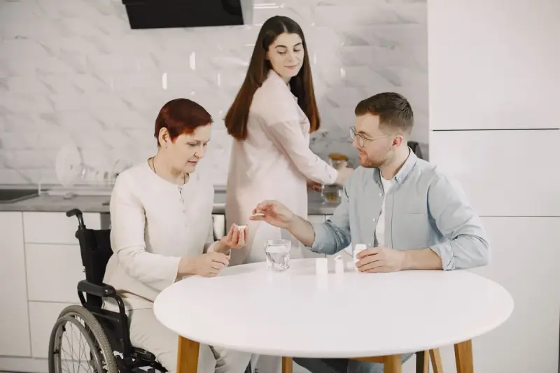 Older woman in a wheelchair at a kitchen table with two adult family members helping her