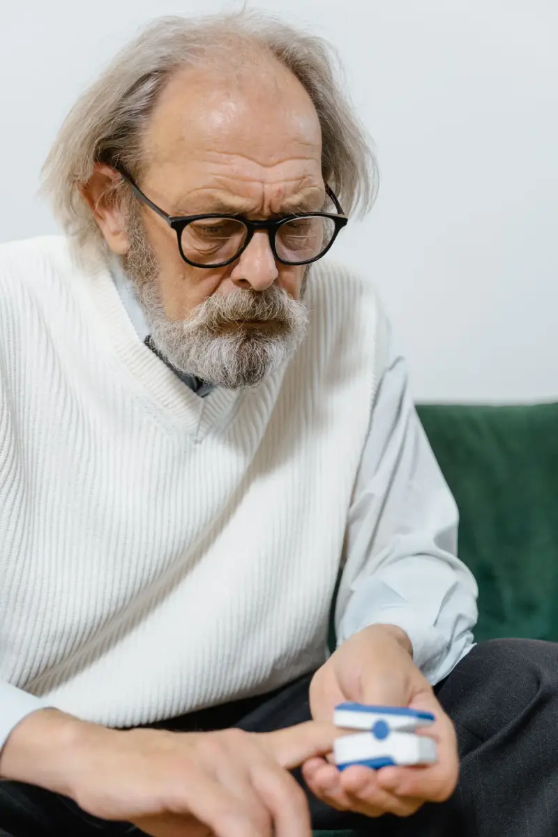 Older man in glasses and a white sweater vest reading a fingertip pulse oximeter at home