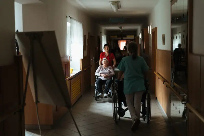 Two caregivers pushing seniors in wheelchairs down a long care home corridor