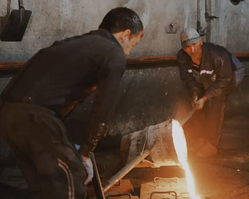 Two foundry workers guiding a hand ladle of molten metal into molds