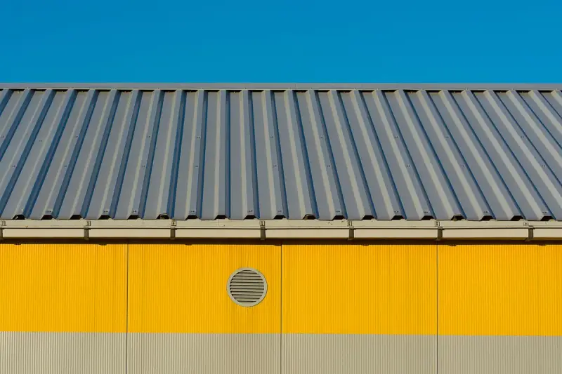 Grey ribbed metal roof above yellow insulated sandwich panel walls with a round vent