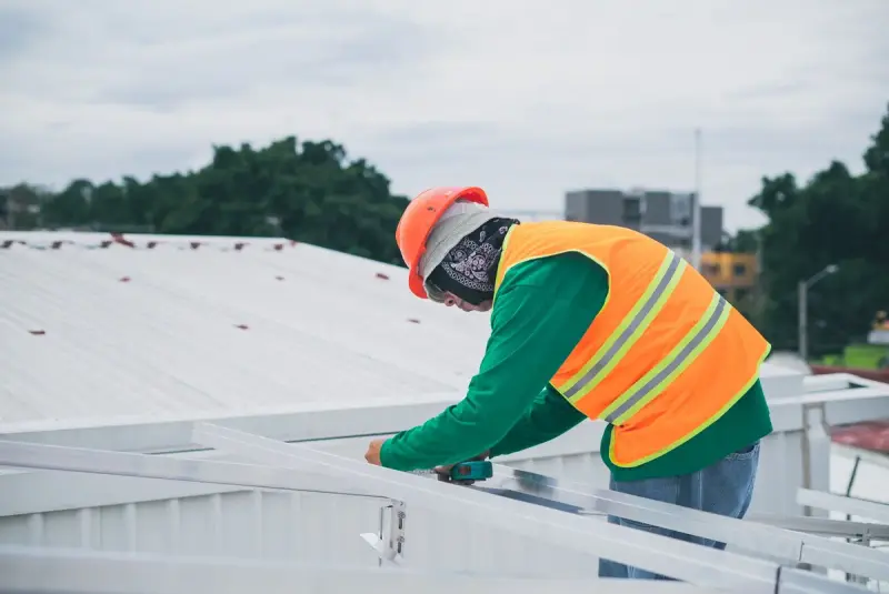 Worker in a hard hat and safety vest fastening aluminum rails on a white metal roof