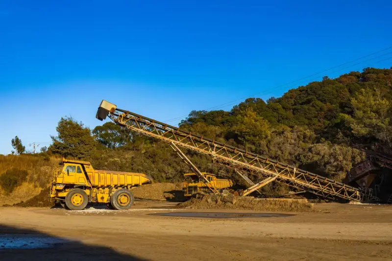 Yellow rigid dump truck beside a long stacking conveyor at a mine site under blue sky