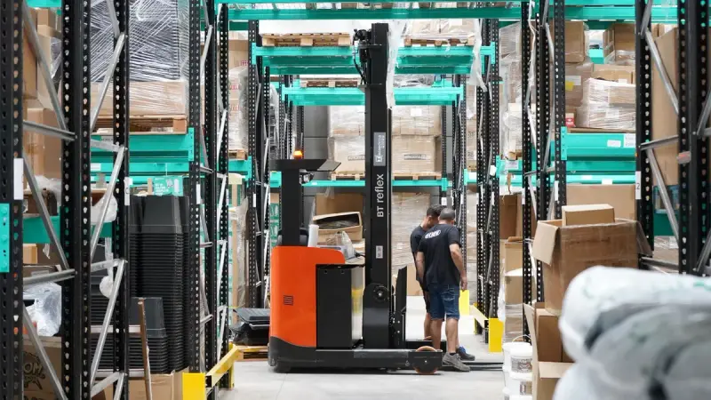 Warehouse workers and a forklift moving through storage aisles