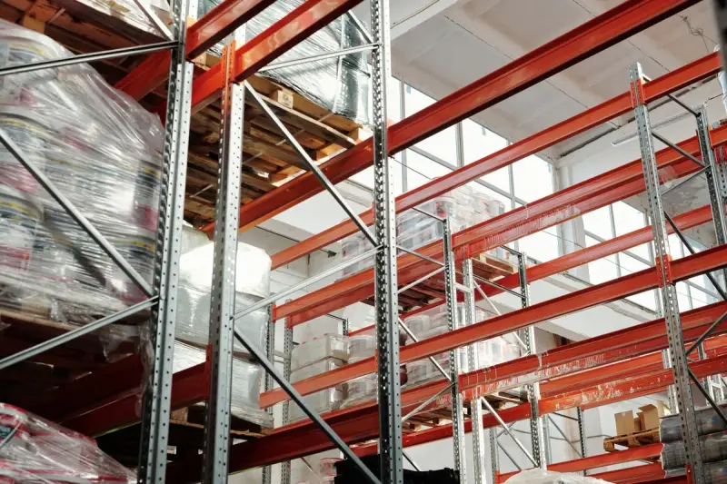 Shrink-wrapped pallets stacked high on orange and steel racking under skylights
