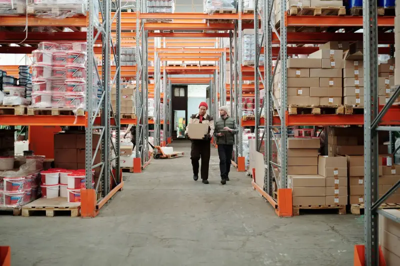 Two warehouse workers carrying a carton along a racked aisle