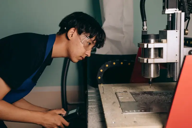 Young machinist in safety glasses leaning in to watch a small CNC mill cut a plate
