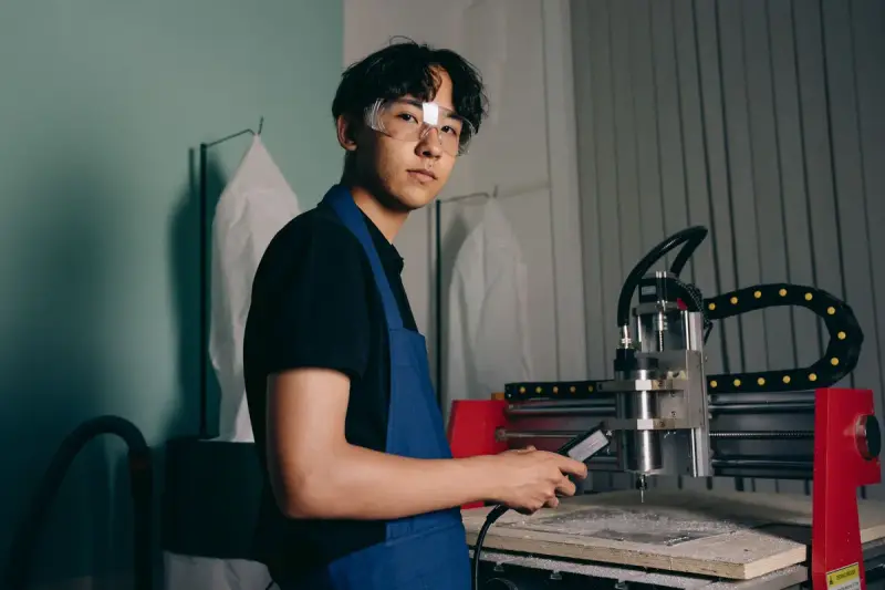 Young engineer in safety glasses and apron holding a handheld controller beside a CNC machine