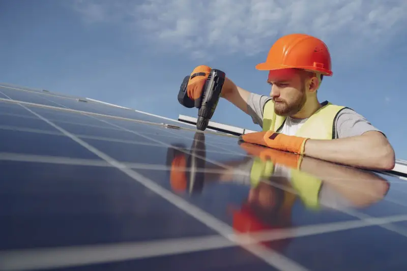 Electrician in a hard hat and gloves drilling while installing a solar panel