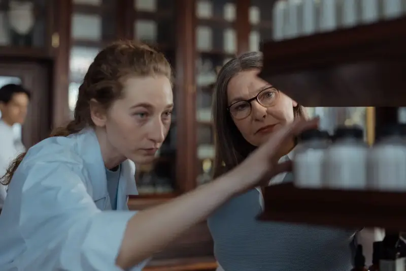 Two pharmacists examining medicine bottles on a wooden pharmacy shelf