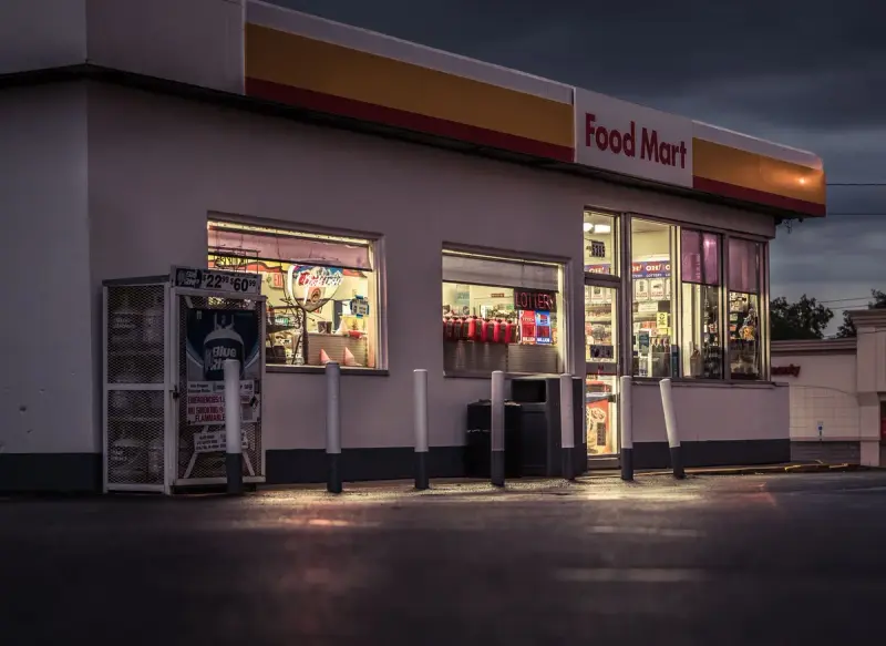 Convenience store food mart at dusk with lit windows, bollards and a propane exchange cage