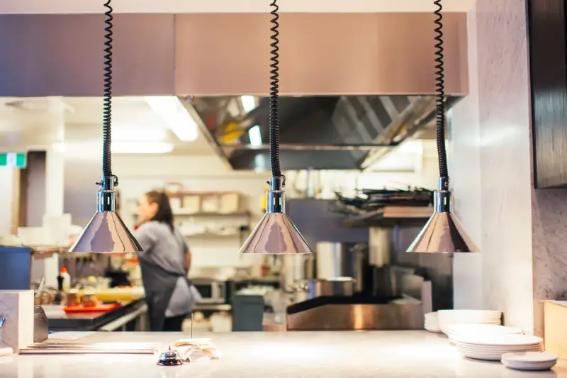 Woman working behind a restaurant kitchen serving counter under hanging lamps