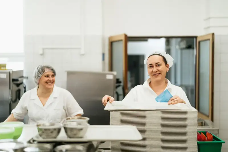 Two smiling kitchen staff in white uniforms and hairnets working in a commercial cafeteria kitchen
