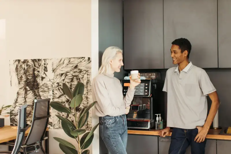 Two coworkers talking over coffee beside an espresso machine in an office pantry