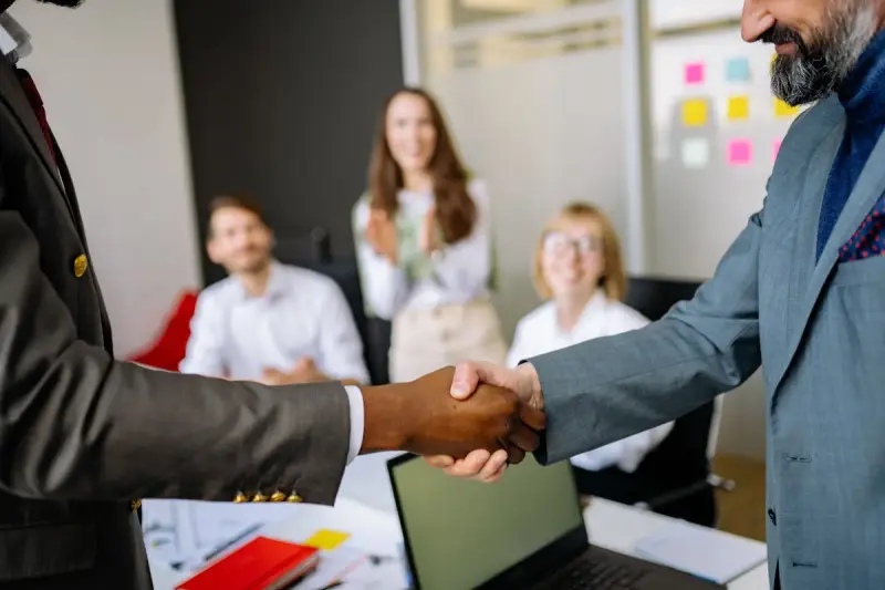 Two business people shaking hands while colleagues applaud in a meeting room