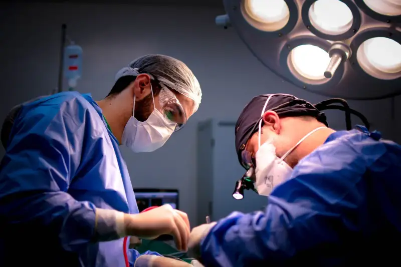 Two surgeons in blue gowns, masks and loupes operating under a bright surgical light
