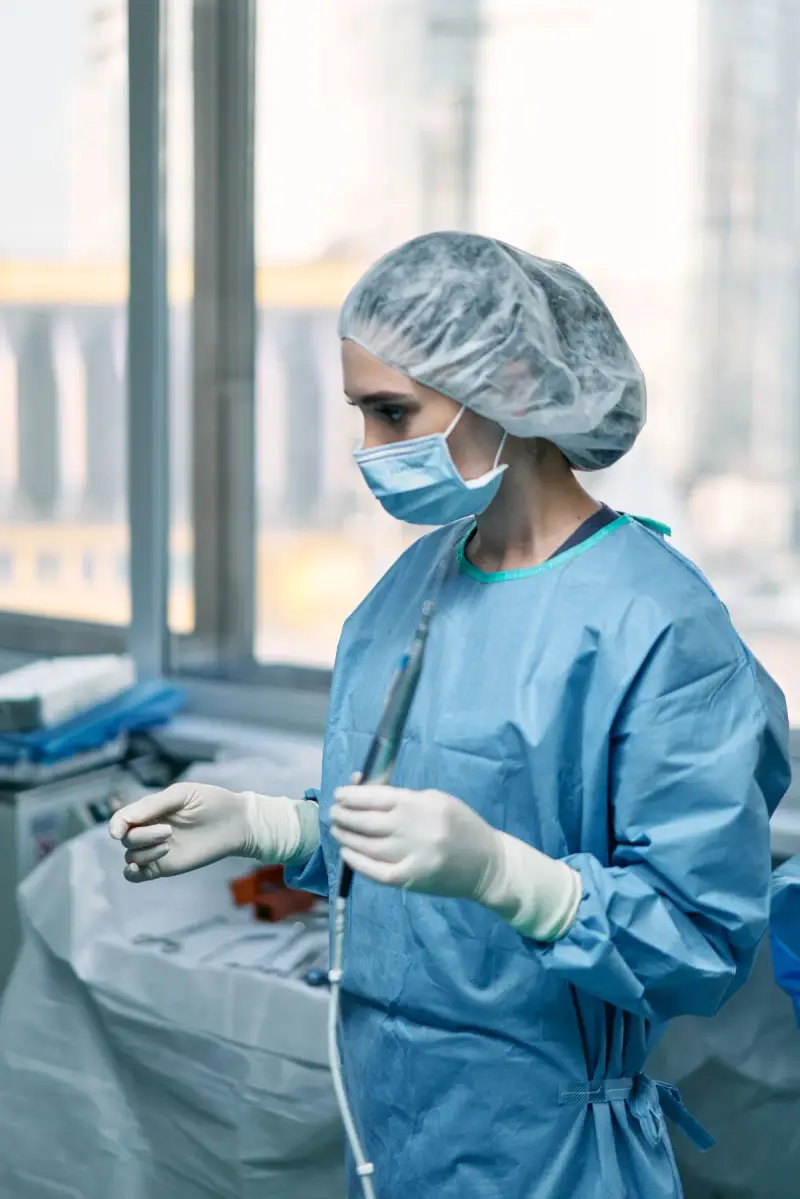 Female surgeon in a blue gown, cap and mask holding a surgical instrument by a window