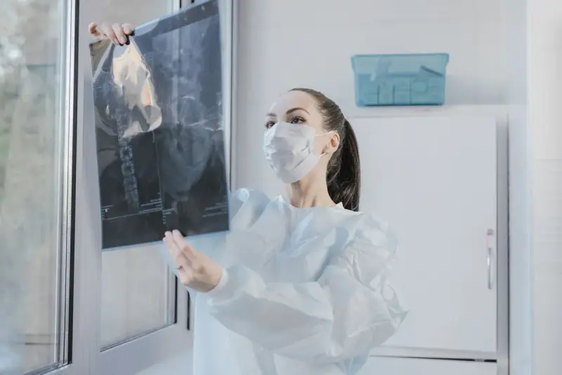 Woman clinician in a mask and gown holding a spine X-ray up to window light