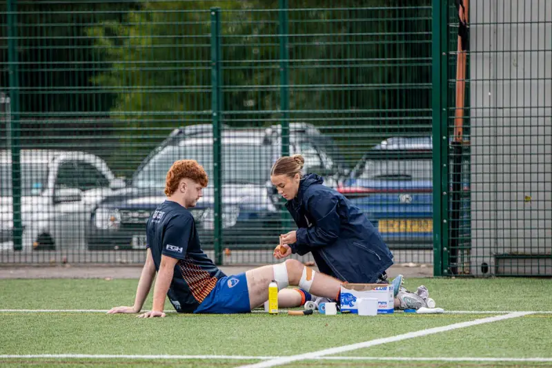 Sports therapist kneeling on artificial turf to tape an injured player's knee beside a kit bag