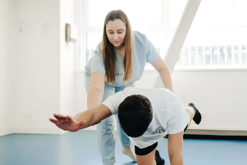 Woman clinician in light blue scrubs guiding a man through a balance exercise on a gym floor