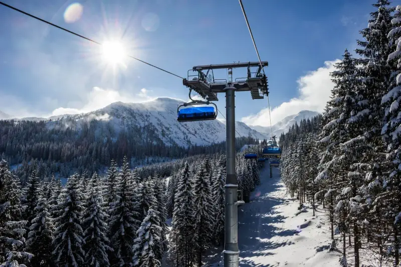Chairlift rising over snowy pine forest toward mountains under a bright winter sun