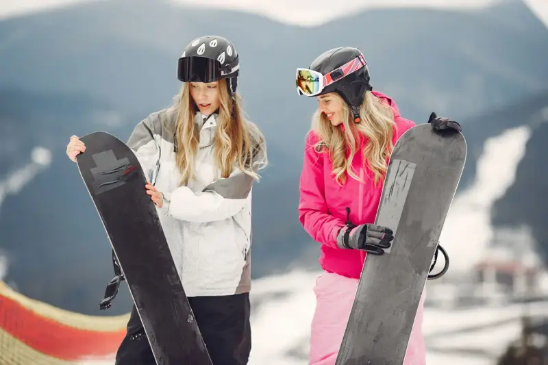 Two women in helmets and goggles holding snowboards on a mountain