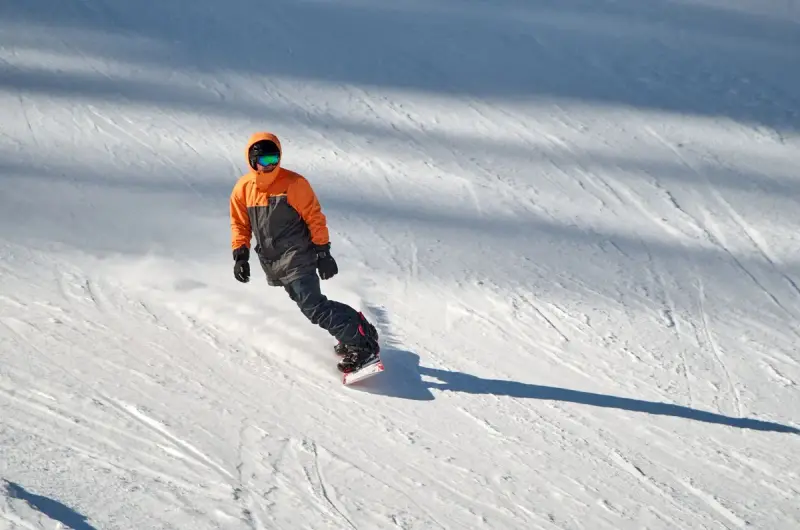Snowboarder in an orange jacket carving across a groomed slope