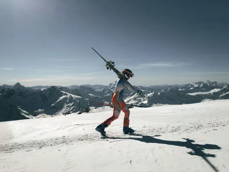 Skier carrying skis over a shoulder across a snowy ridge with peaks behind