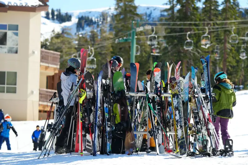 Skiers and snowboarders gathered by crowded gear racks at a sunny mountain resort