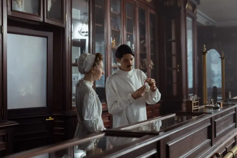 Two pharmacists in white gowns behind a glass-topped counter examining a small medicine bottle