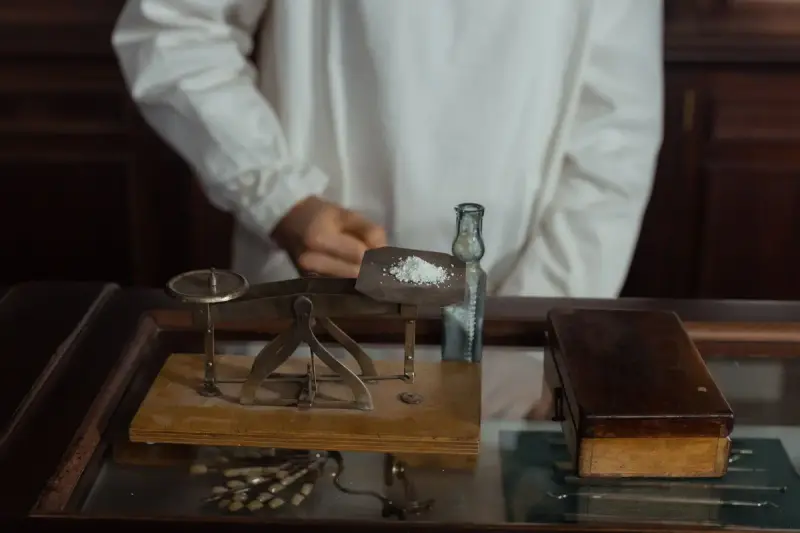 Pharmacist weighing white powder on a brass balance scale beside a glass medicine bottle