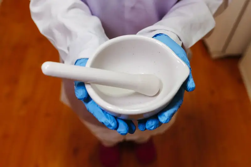 Gloved hands holding a white porcelain mortar and pestle above a wooden counter