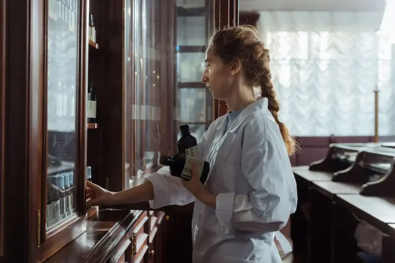 Female pharmacist with a braid taking bottles from a tall glass-fronted medicine cabinet