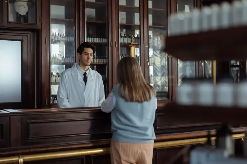 Pharmacist in a white coat and tie listening to a customer at a wooden pharmacy counter